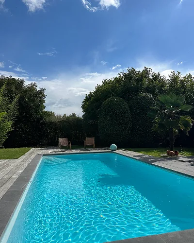 A turquoise swimming pool with a wooden deck, in a garden surrounded by tall hedges
