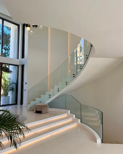 A bright villa hallway with a curved staircase, glass balustrade and lit steps